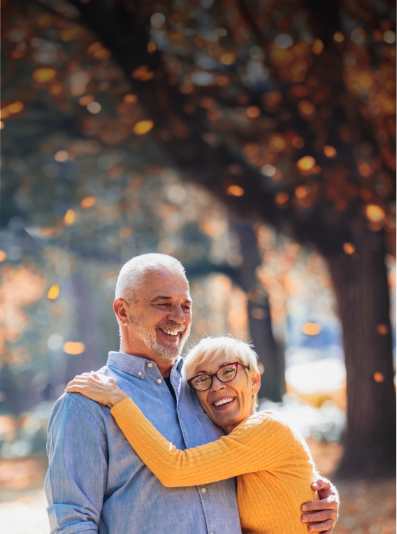 Older man and woman hugging outdoors with autumn trees in background